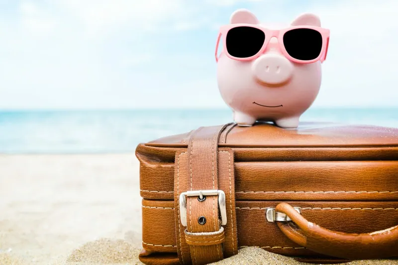 Sparschwein mit Sonnenbrille steht auf einem Reisekoffer am Strand